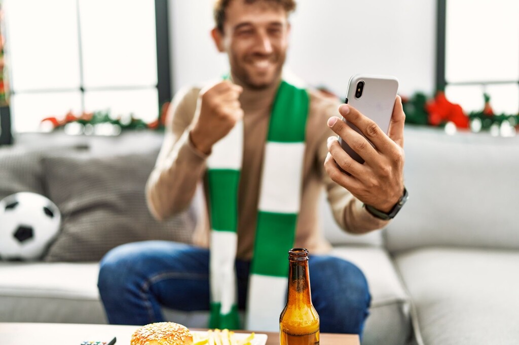 Homem de camisa marrom e cachecol branco e verde, olhando um celular e comemorando sentado em um sofá.