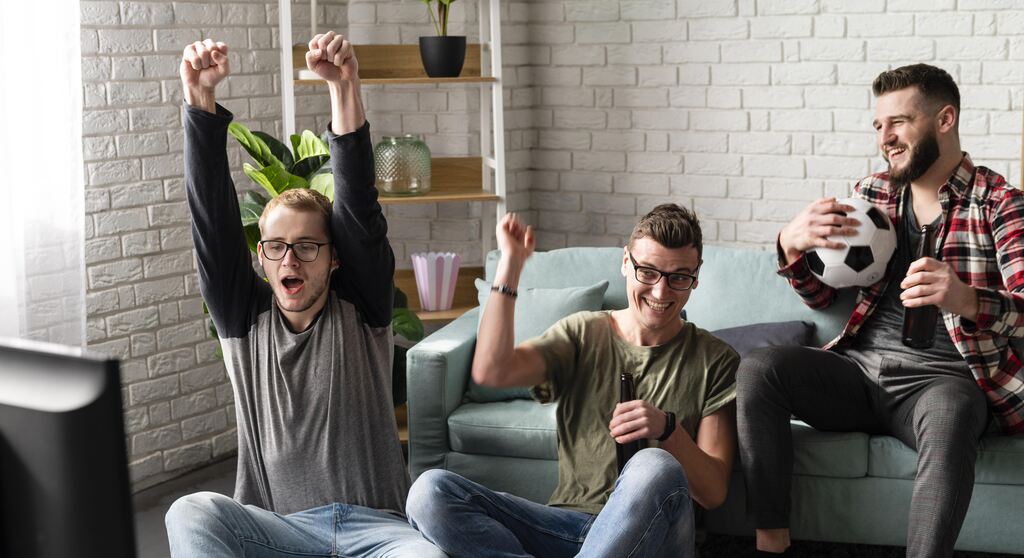 3 homnes emuma sala com sofá azul, sentados, tomando cerveja e comemorando enquanto assistem TV.