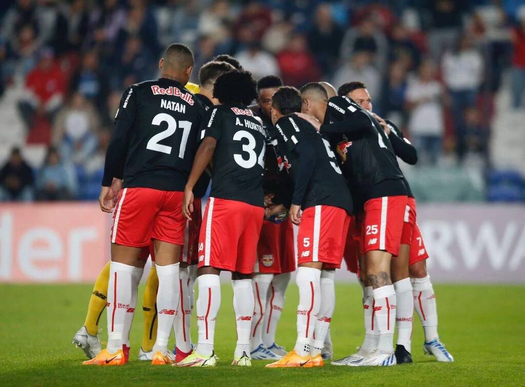 Jogadores do Red Bull Bragantino vestindo uniforme vermelho e preto, abraçados, reunidos em círculo no gramado durante jogo.