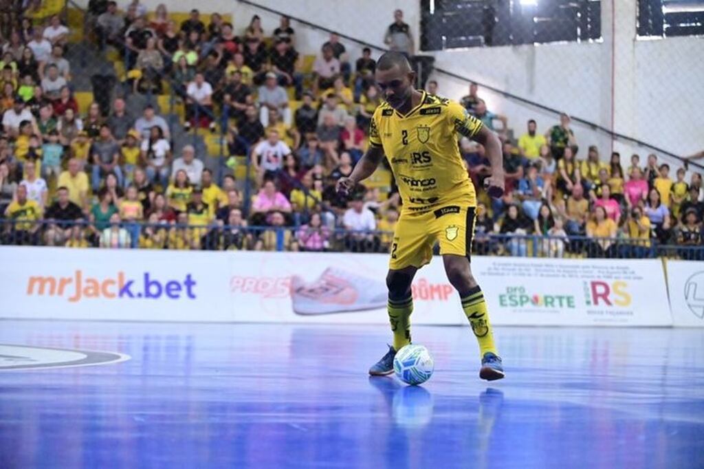 Jogador de futsal vestindo uniforme amarelo, conduzindo a bola pela quadra durante a partida.