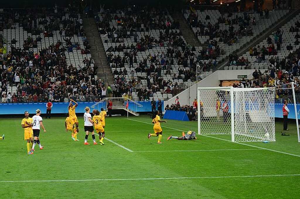 Jogadoras de futebol feminino, vestindo uniforme amarelo e outras de uniforme branco, dentro da grande área, com bola no gol.