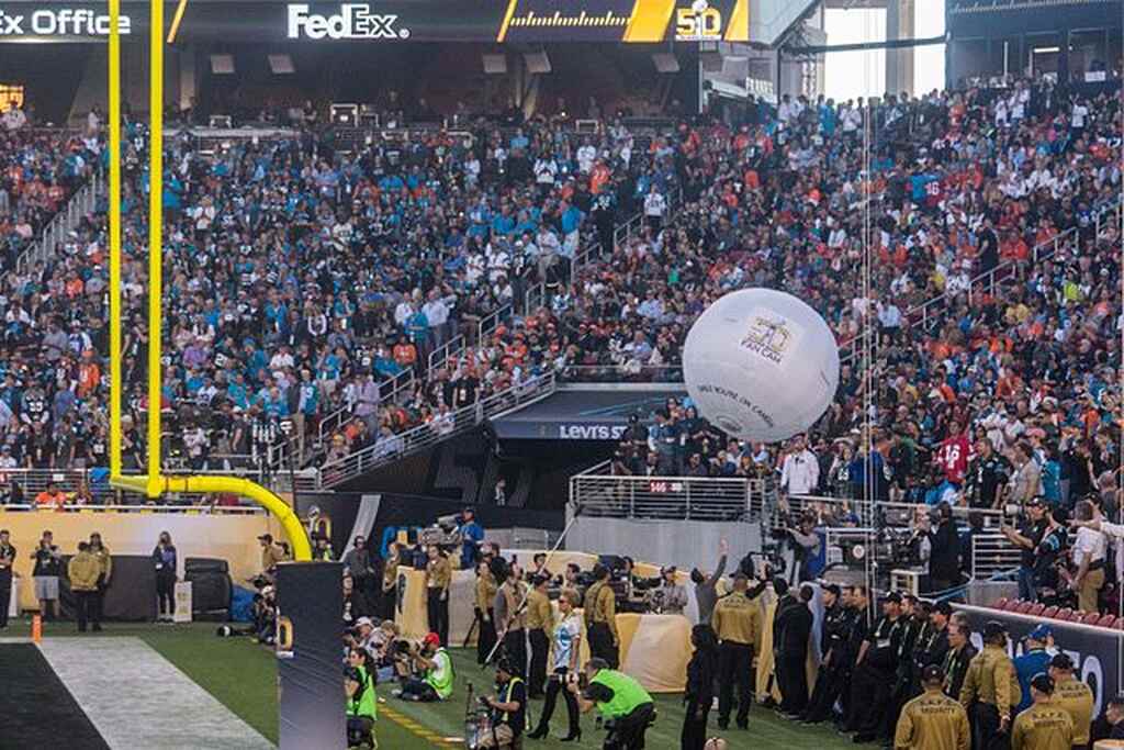 Arquibancadas cheias de torcedores vendo uma partida de futebol americano e muitos trabalhadores do evento na parte de baixo.