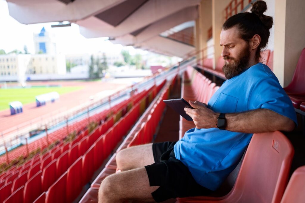 Homem de barba usando camisa azul e segurando um tablet, pensativo, sentado em cadeiras vermelhas de uma arquibancada.