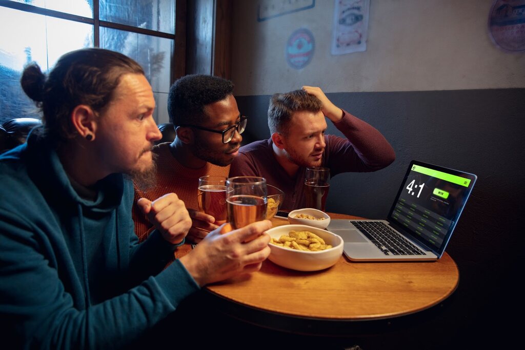 3 homens sentados em uma mesa, tomando cerveja e comendo salgadinhos enquanto acompanham um placar pelo notebook.