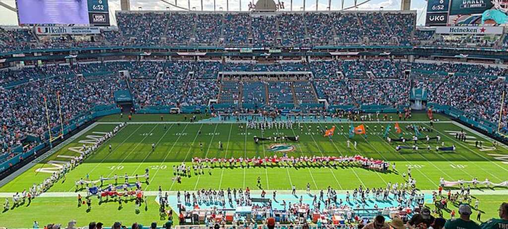 Estádio de futebol americano lotado, com arquibancadas cheias durante partida, com jogadores andando pelo gramado.