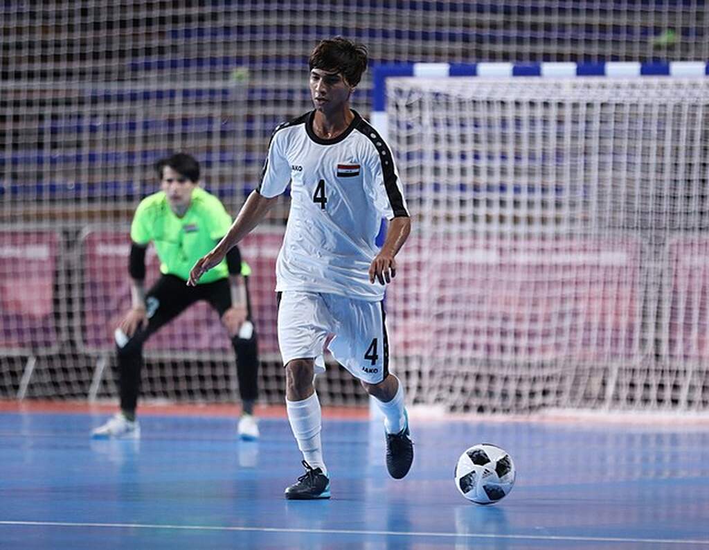 Jogador de futsal vestindo uniforme branco com listra preta, conduzindo uma bola ao longo de uma quadra azul.