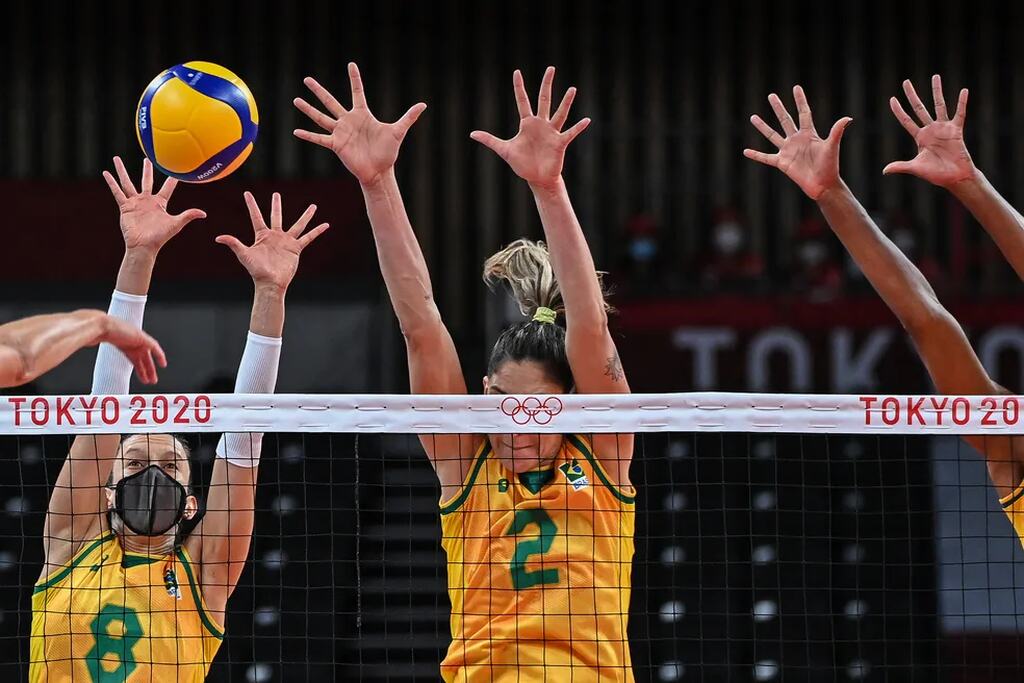 Jogadpras de vôlei vestindo uniforme da seleção brasileira realizando bloquei na rede durante partida profissional.