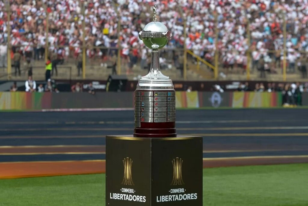 Taça da Libertadores colocada sobre suporte no meio de estadio de futebol, com a torcida ao redor.