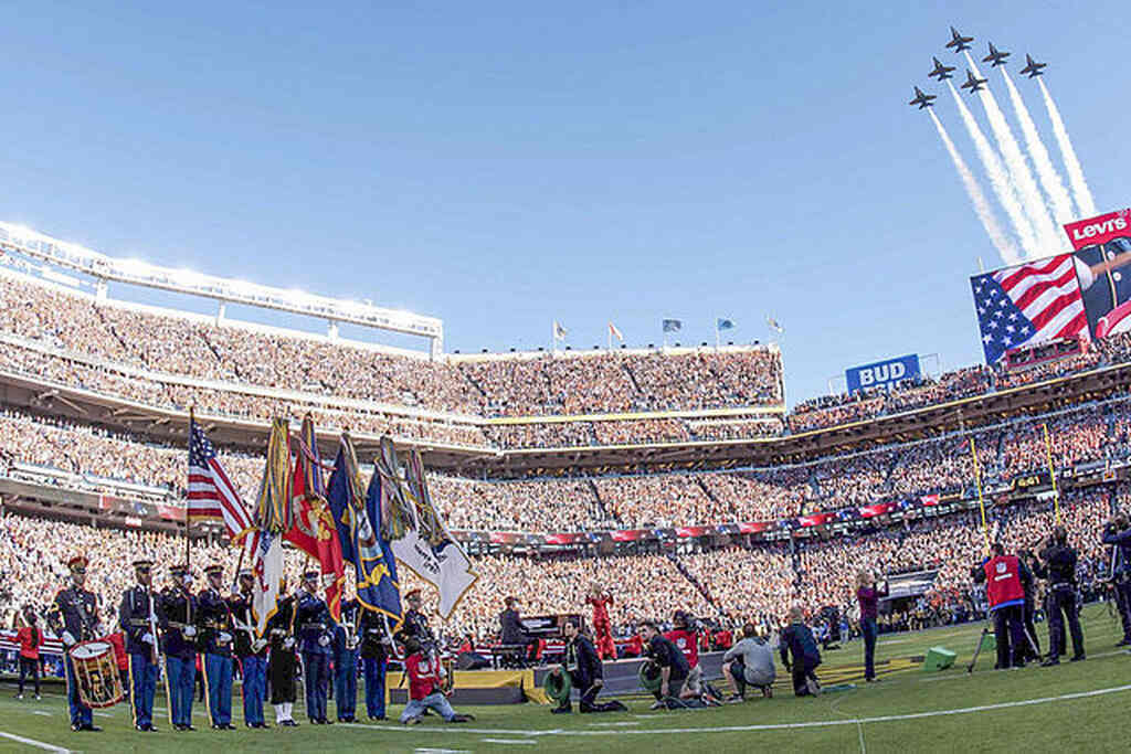 Arquibancadas de estádio de futebol americano lotadas, durante Super Bowl com banda posicionada no meio do gramado.