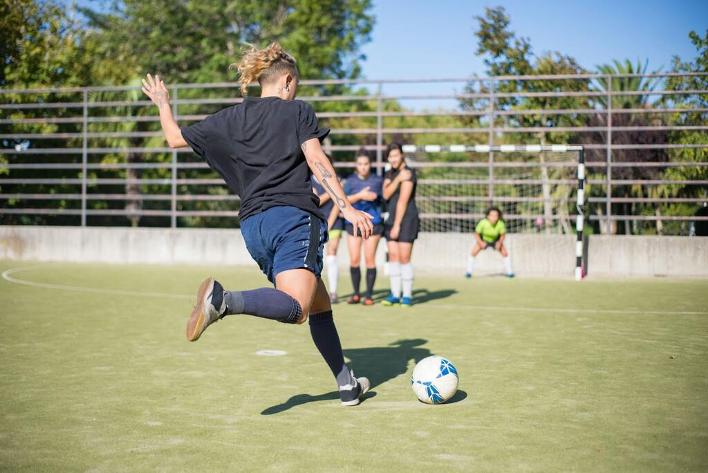 Jogadora de futebol realizando uma cobrança de falta contra o gol, com 3 jogadoras na barreira.