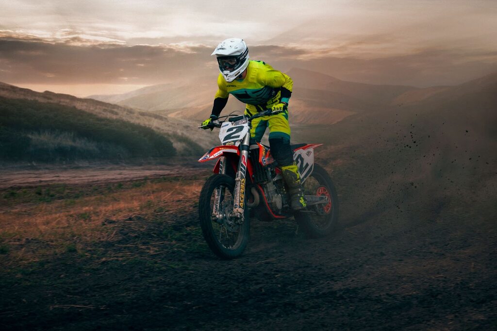 Piloto de moto vermelha, vestindo uniforme amarelo e verde e usando capacete em pista de terra.