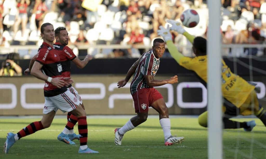 Jogador do Fluminense finalizando contra o Gol enquanto goleiro faz a defesa e marcador do Flamengo fica observando.