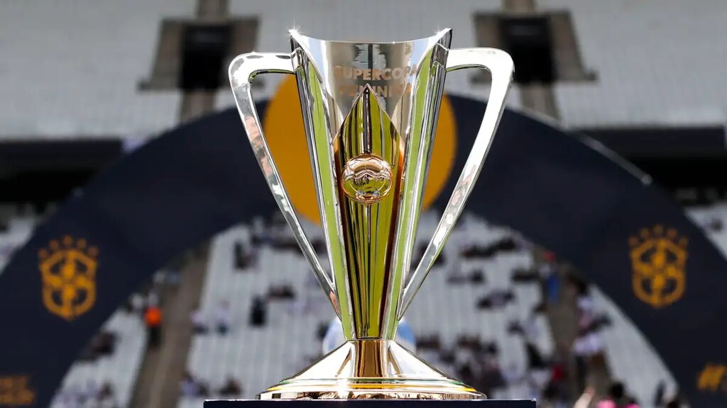 Taça dourada da Supercopa feminina de futebol, colocada em cima de um pedestal em um estádio.