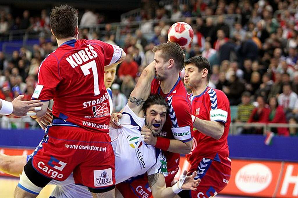 Jogador profissional de handebol, durante partida, vestindo uniforme branco e sendo marcado por 3 jogadores de vermelho.