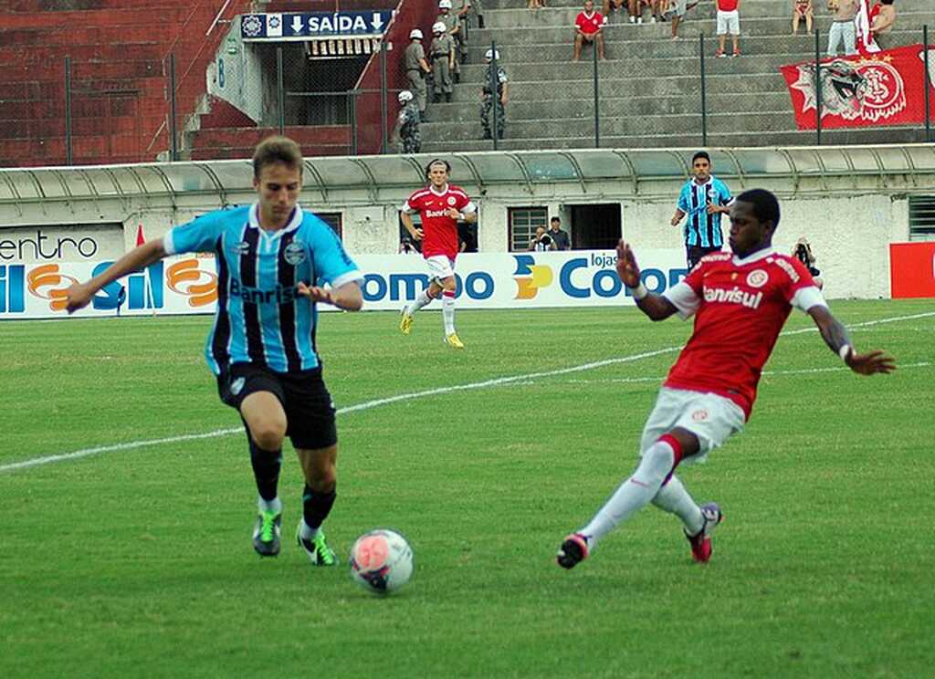 Jogador do Internacional, de uniforme vermelho e branco, tenta roubar a bola do jogador do Grêmio, de uniforme azul e preto.