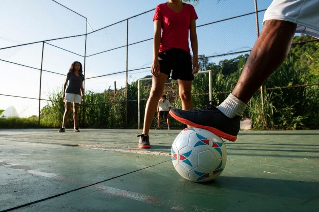 Homem pisando na bola em quadra de futsal, enquanto joga com outro homem e duas mulheres.