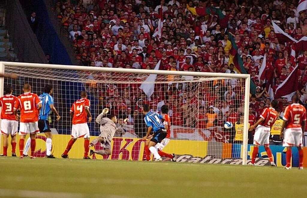 Jogador do Grêmio marca um gol contra o Internacional. Ao fundo, está a torcida colorada nas arquibancadas.