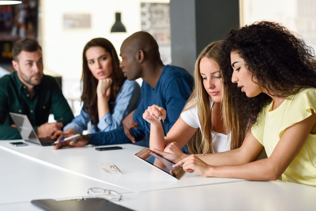 Três mulheres e dois homens durante uma reunião. Há celulares e notebooks em cima da mesa.