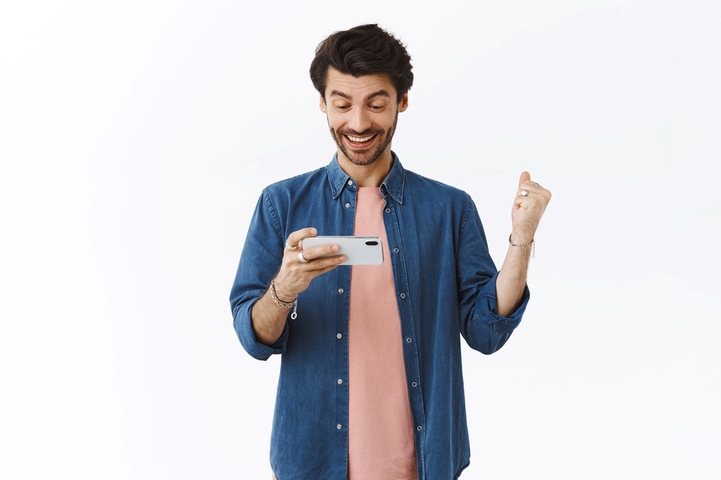 Homem de camiseta rosa e camisa jeans aberta, comemorando com expressão de felicidade, enquanto segura um smartphone.