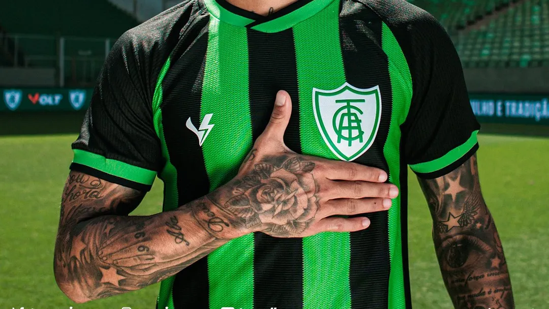 Jogador do América-MG vestindo camisa verde e preta do clube, com a mão no peito.