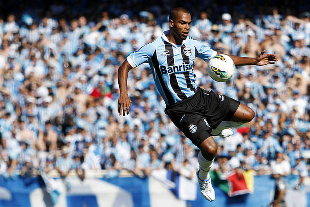 Jogador do Grêmio, de uniforme azul e preto, domina a bola. Ao fundo, está a torcida gremista nas arquibancadas.