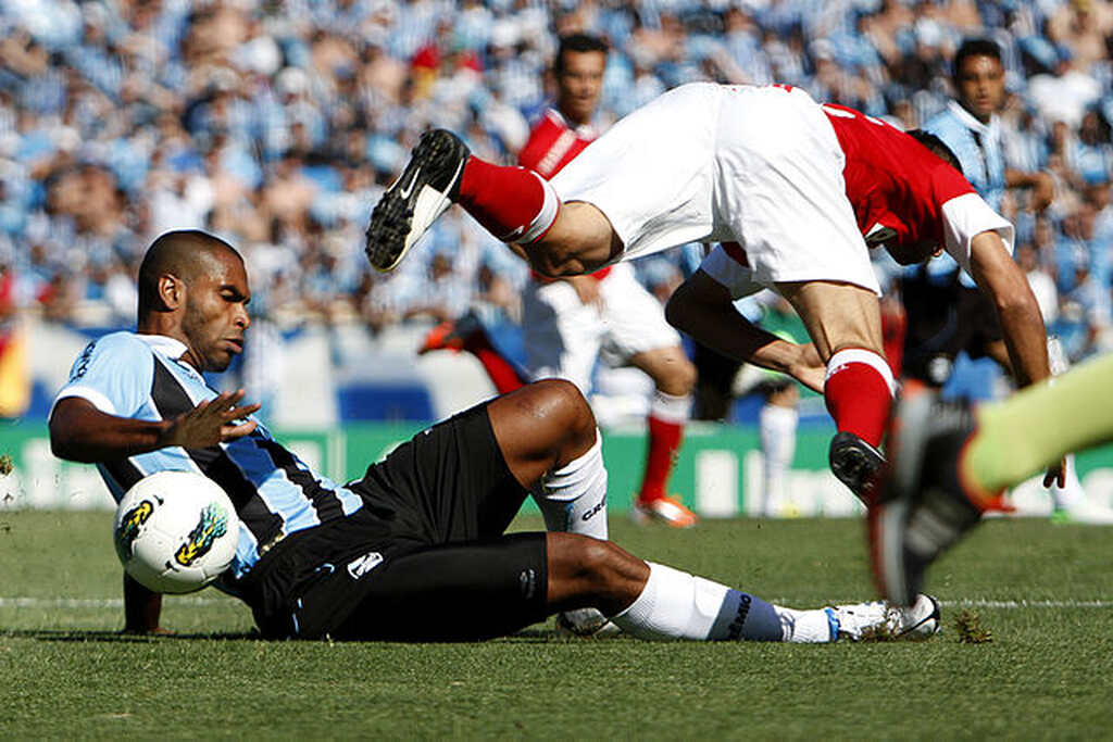 Jogador do Grêmio, de uniforme azul e preto, aplica carrinho no adversário do Internacional, de uniforme vermelho e branco.