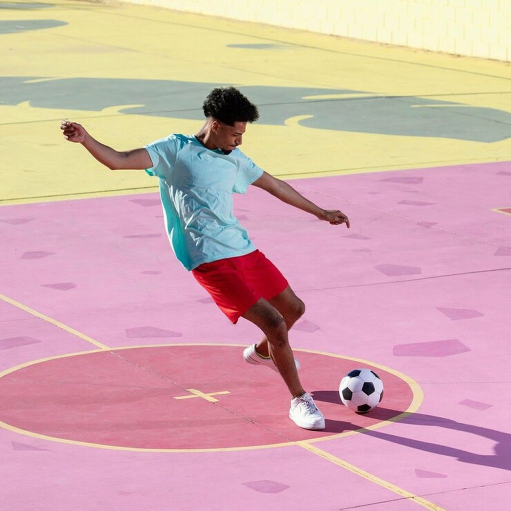 Homem com uma camiseta azul claro e um calção vermelho, fazendo o movimento para chutar a bola em uma quadra de futsal.