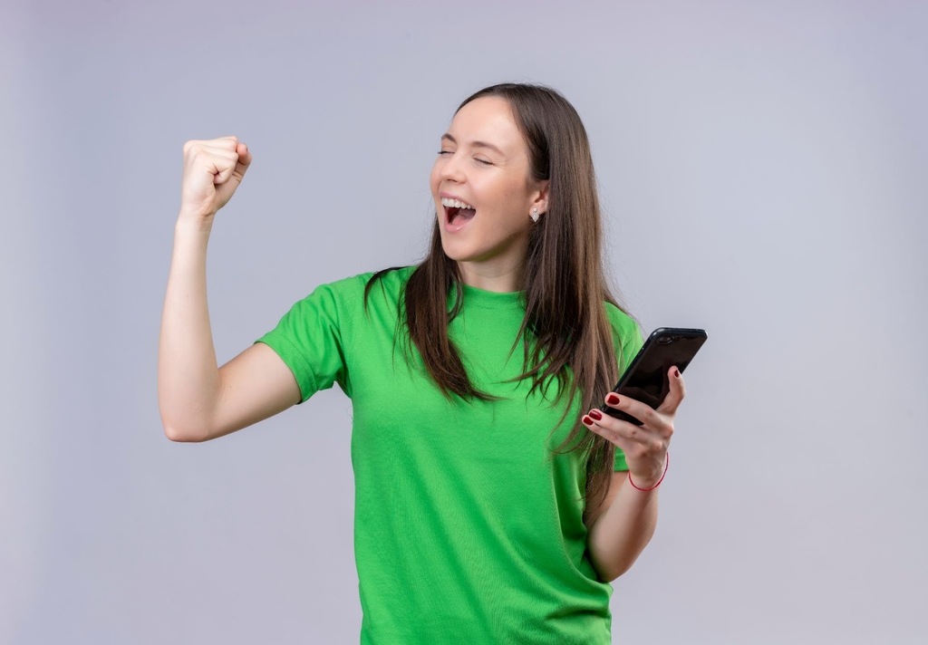 Mulher de camiseta verde comemorando, com expressão de felicidade e punho cerrado, enquanto segura um smartphone.