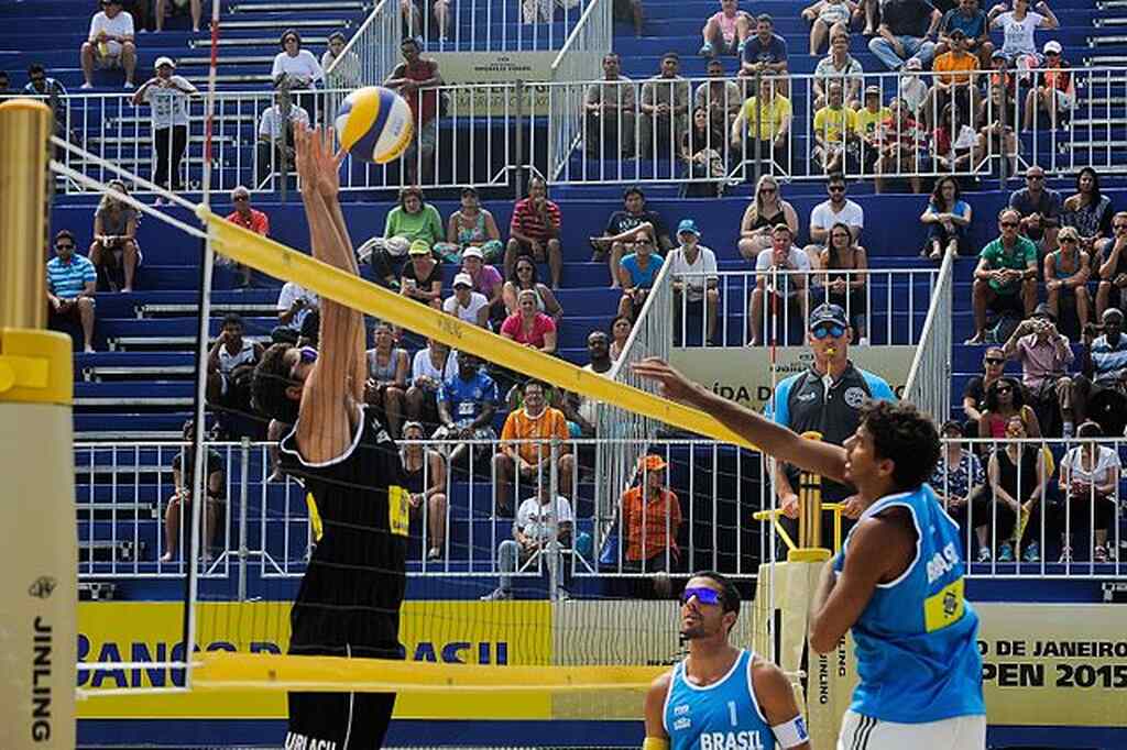 Jogador de vôlei de praia vestindo uniforme preto, bloqueando cortada de jogador de uniforme azul.