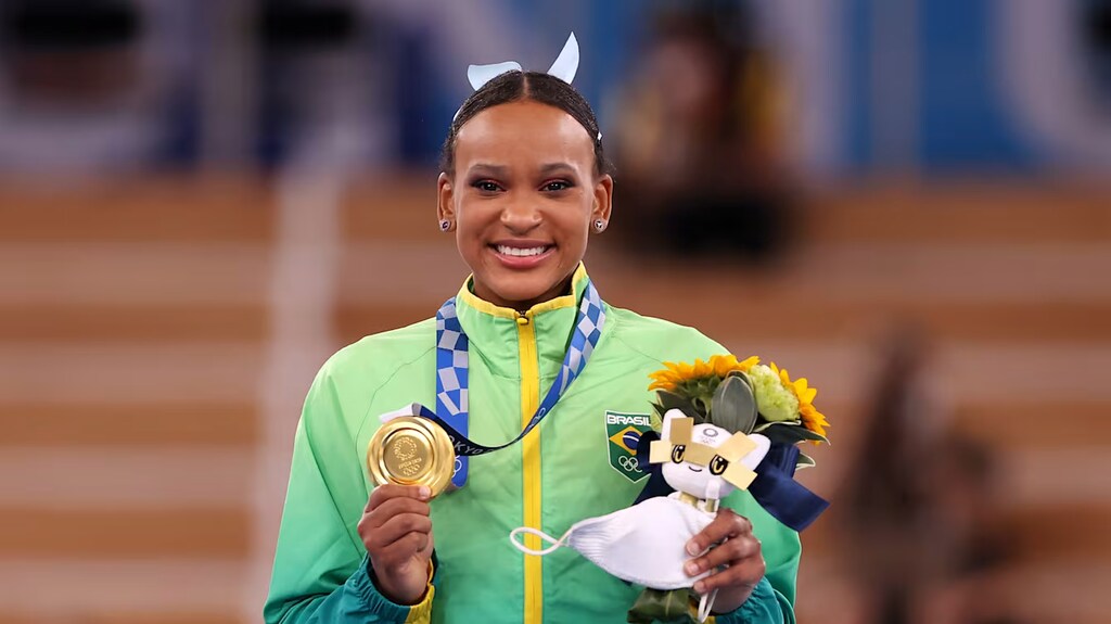 Atleta olímpica brasileira, Rebeca Andrade, exibindo medalha de ouro com a mão direita, enquanto segura flores com a mão esquerda.