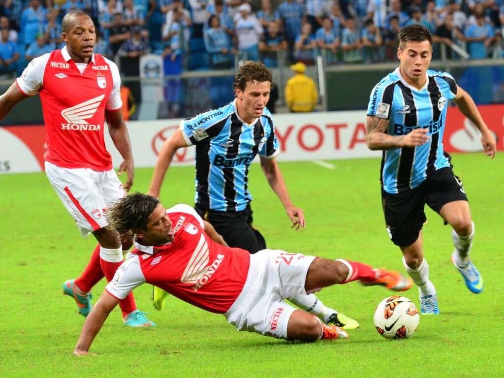 Jogadores do Grêmio, de uniforme azul com listras pretas, driblando jogador de uniforme vermelho e branco.