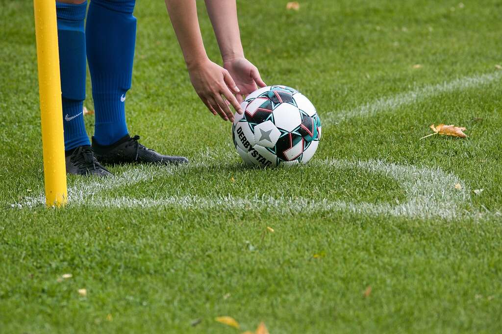 Jogador de futebol usando chuteira preta e meião azul, ajustando a bola na marca de escanteio na linha de fundo.