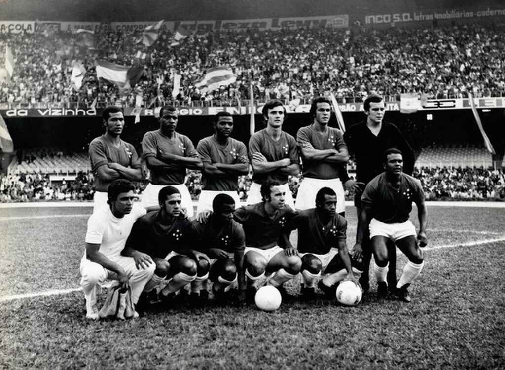 Jogadores do Cruzeiro em campeonato antigo, vestindo calção branco, no meio do gramado, durante partida em preto e branco.