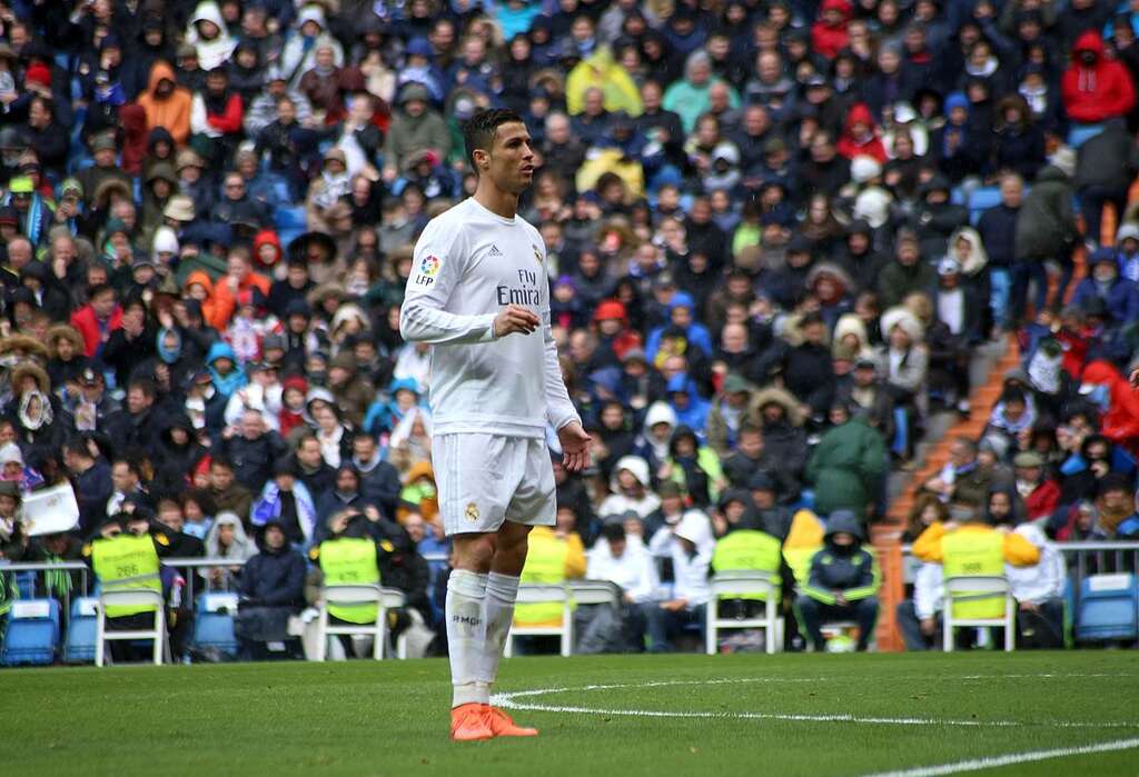 Jogador de futebol Cristiano Ronaldo, com uniforme branco do Real Madrid, comemorando gol diante de torcida adversária.