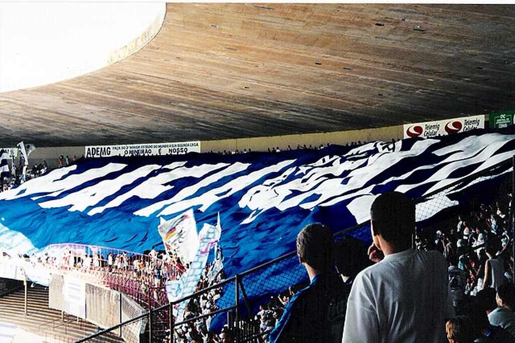 Arquibancada de estádio de futebol, lotada, com torcida do Cruzeiro, com grande bandeira azul sobre a torcida.