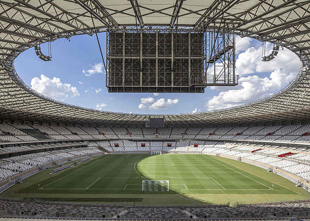 Campo de futebol visto do alto do estádio, com arquibancadas brancas, totalmente vazias.