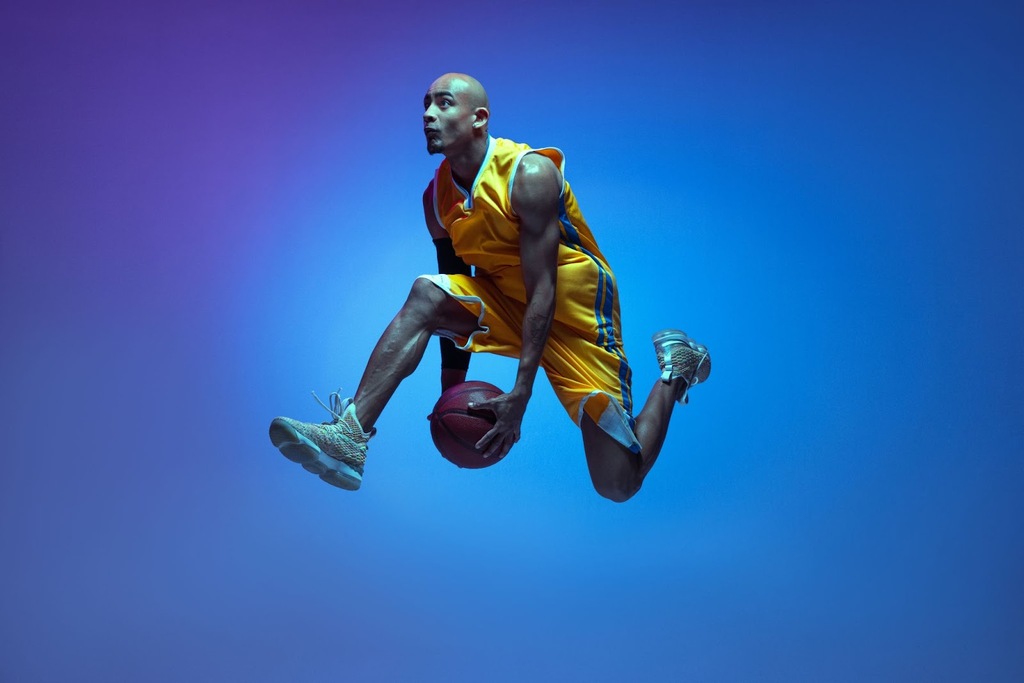 Jogador de basquete de uniforme amarelo, com tênis branco, segurando uma bola de basquete de baixo das pernas em fundo azul.