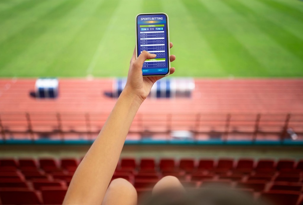 Pessoa segurando um celular na arquibancada de um estádio de futebol, com site de apostas esportivas na tela.