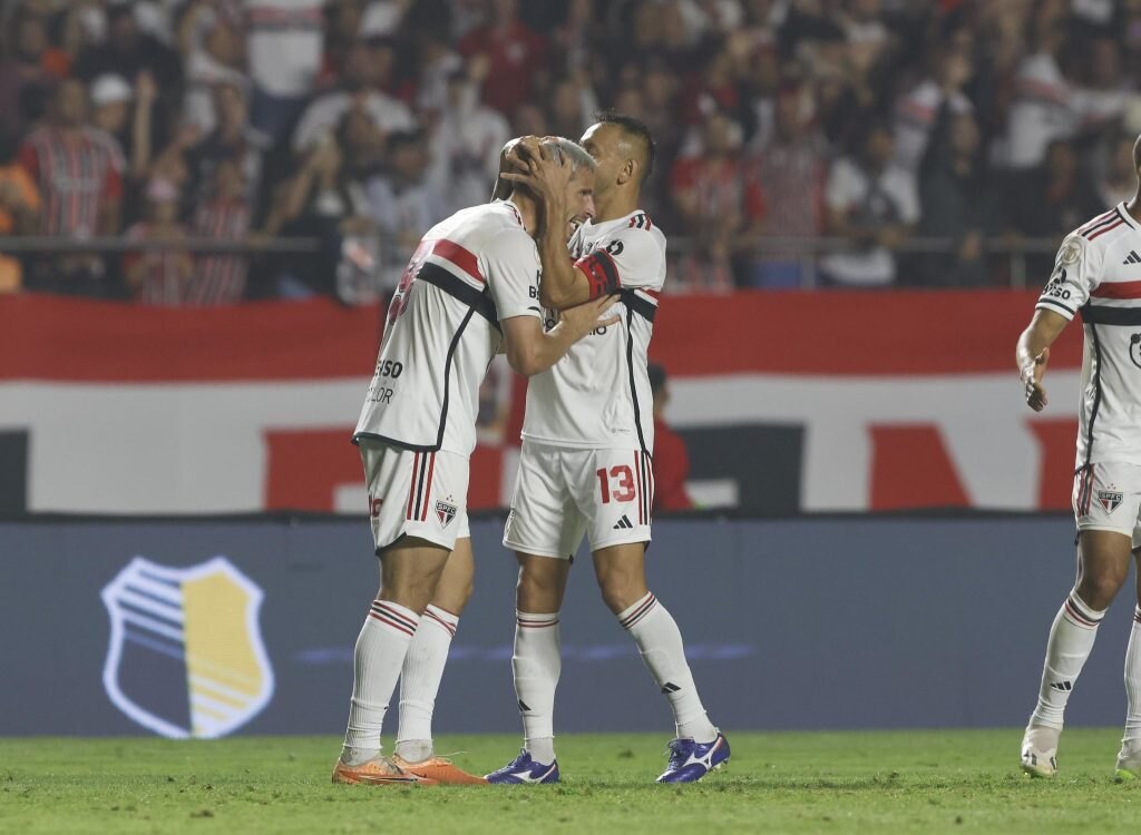 Dois jogadores do São Paulo se abraçando, com uniforme branco com detalhes vermelhos e pretos, durante jogo.