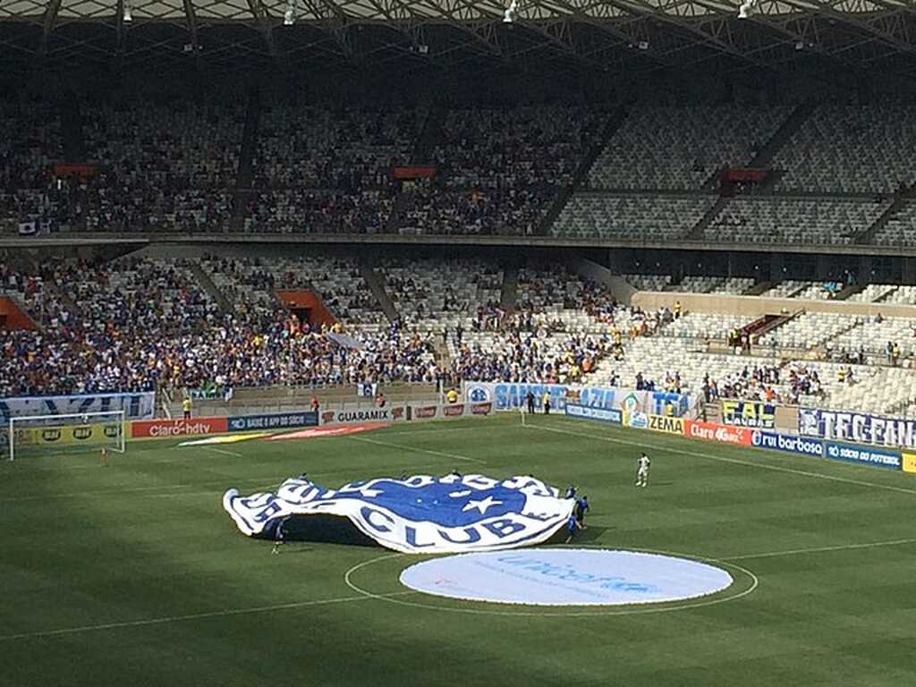 Gramado com pessoas segurando escudo do Cruzeiro na região central, e arquibancadas enchendo.