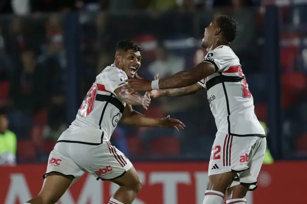 Jogadores do São Paulo vestindo uniforme branco com detalhes vermelhos e pretos, correndo para comemorar gol.