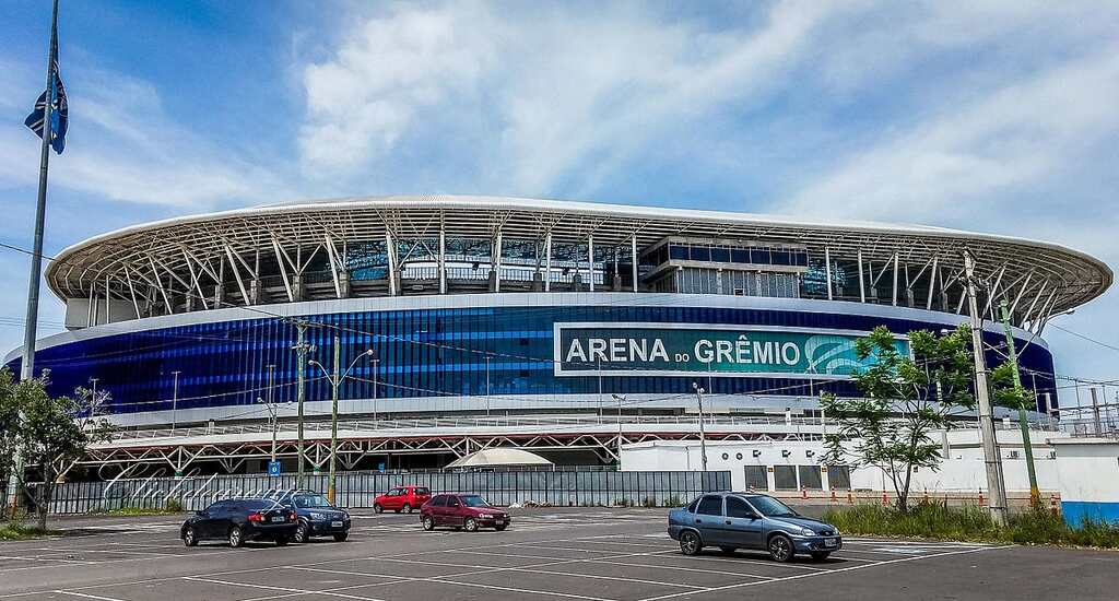 Estádio do Grêmio visto da rua, com carros no estacionamento durante dia de sol.