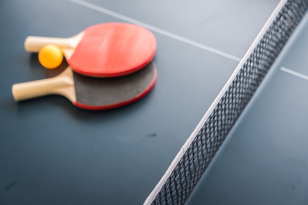 Duas raquetes de tênis de mesa, com borracha vermelha e preta, colocadas em mesa de ping-pong azul, com uma bolinha laranja.