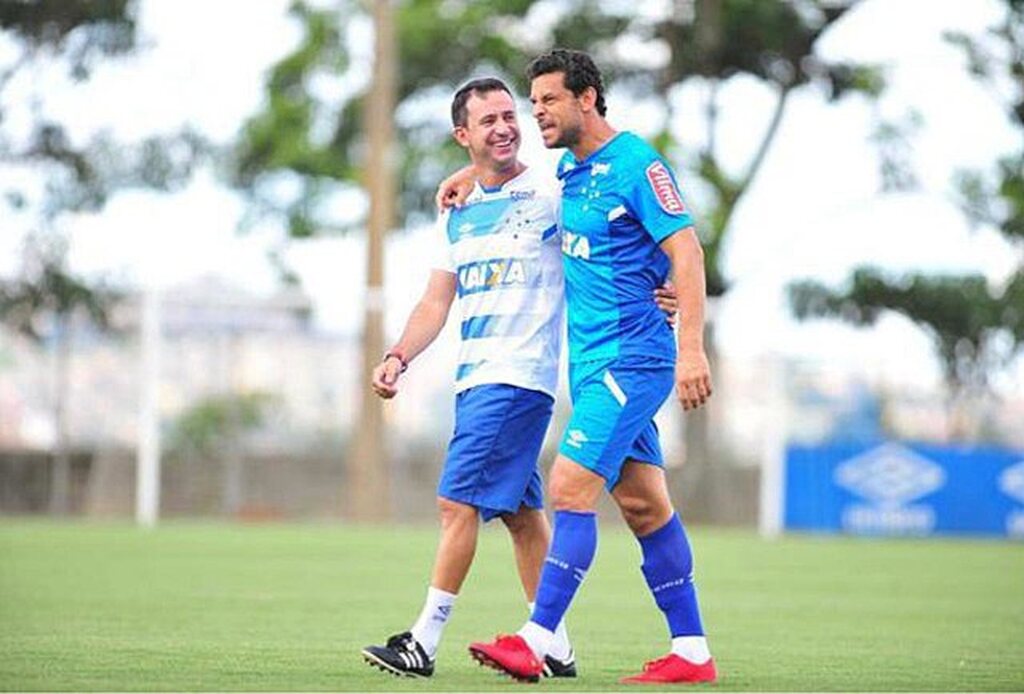 Ex-jogador de futebol, Fred, vestindo uniforme azul do Cruzeiro, abraçado com membro da comissão técnica do clube.