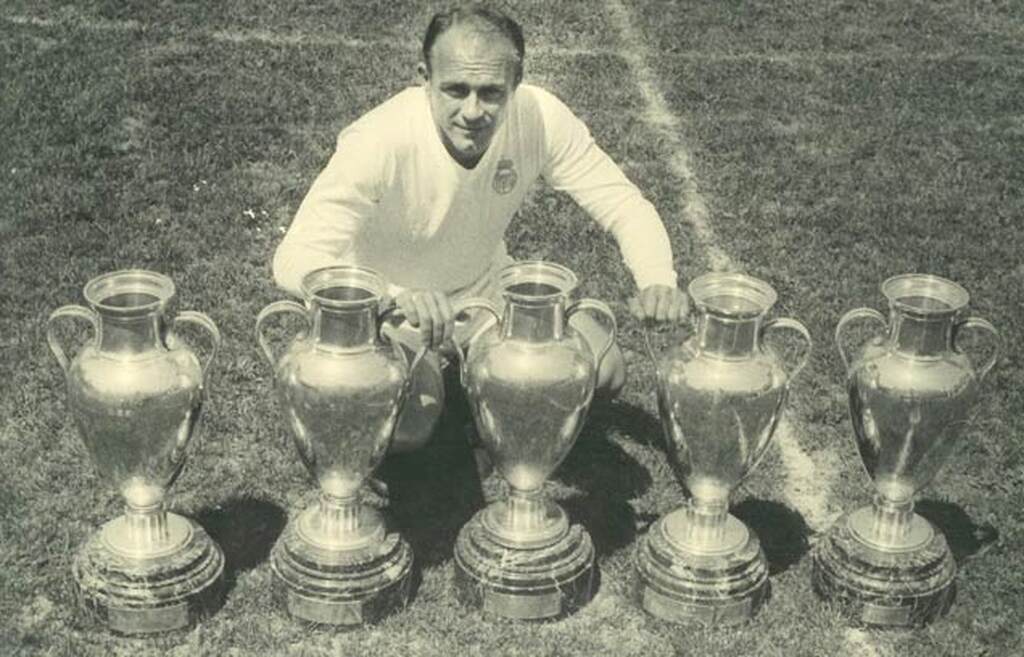 Ex-jogador de futebol, Di Stéfano, agachado no gramado atrás de 5 taças da Champions League.