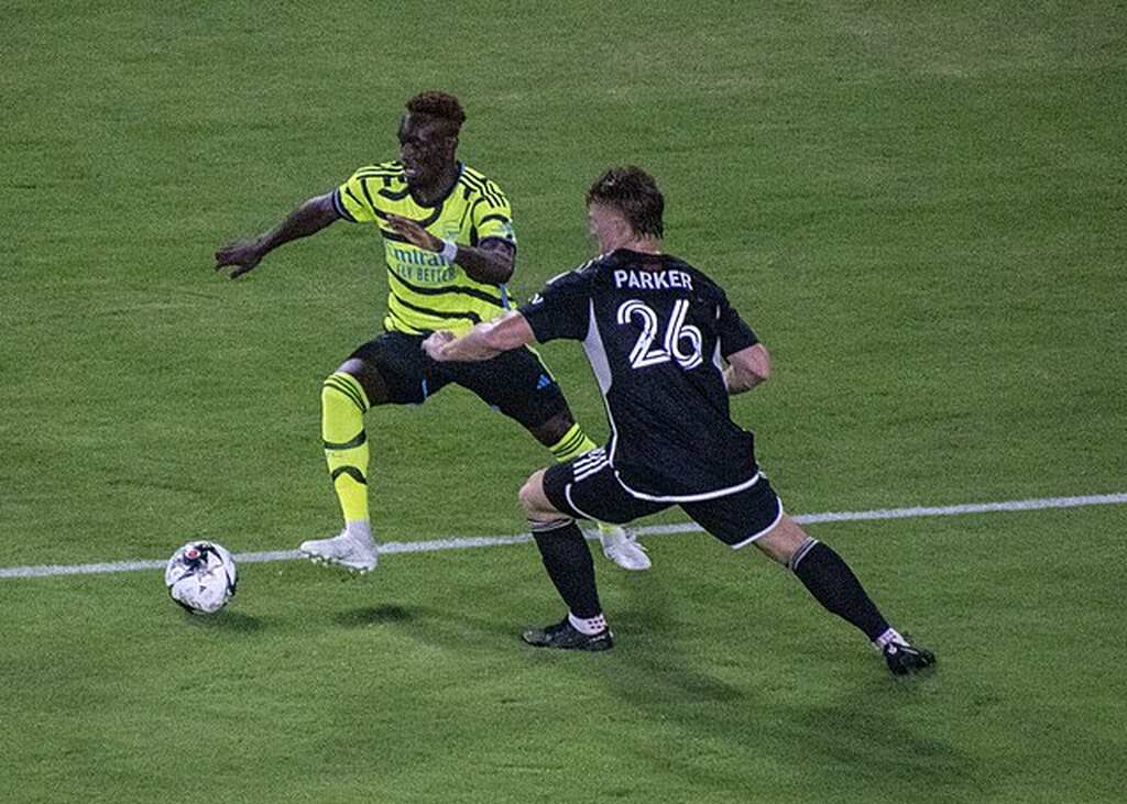 Jogador de uniforme amarelo com listras pretas tentando driblar jogador de uniforme preto durante jogo da MLS.