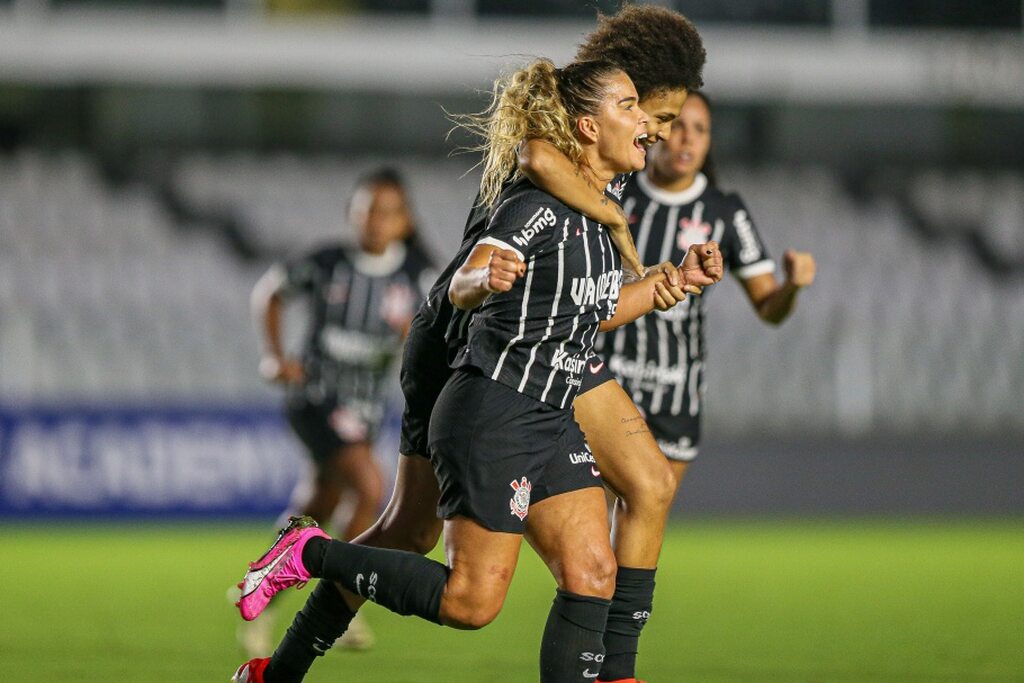 Duas jogadoras do Corinthians, vestindo uniforme preto com listras brancas, comemorando gol durante partida.