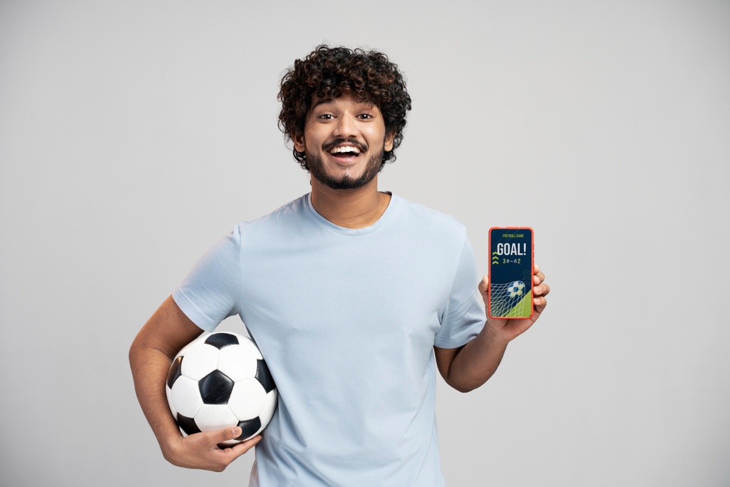 Homem de camisa branca, segurando bola de futebol com o braço direito e mostrando um celular com gol na tela.