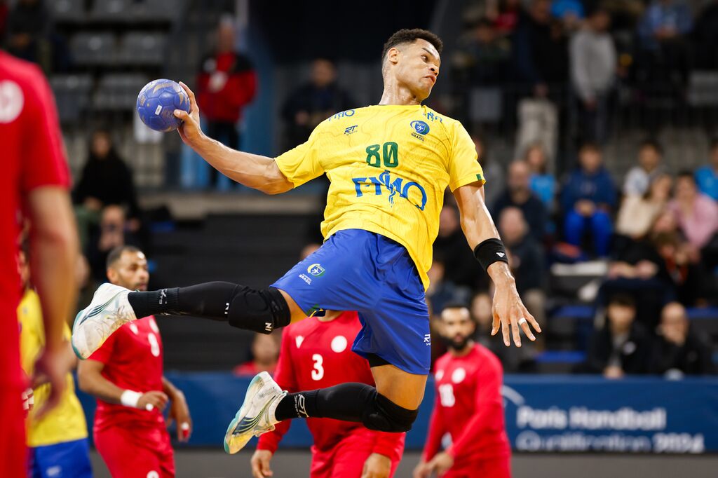 JOgador de handebol da seleção brasileira, usando uniforme amarelo e azul, no ar, arremessando.