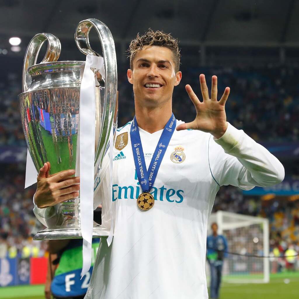 Jogador de futebol, Cristiano Ronaldo, vestindo uniforme branco do Real Madrid, segurando a taça da Champions e comemorando.